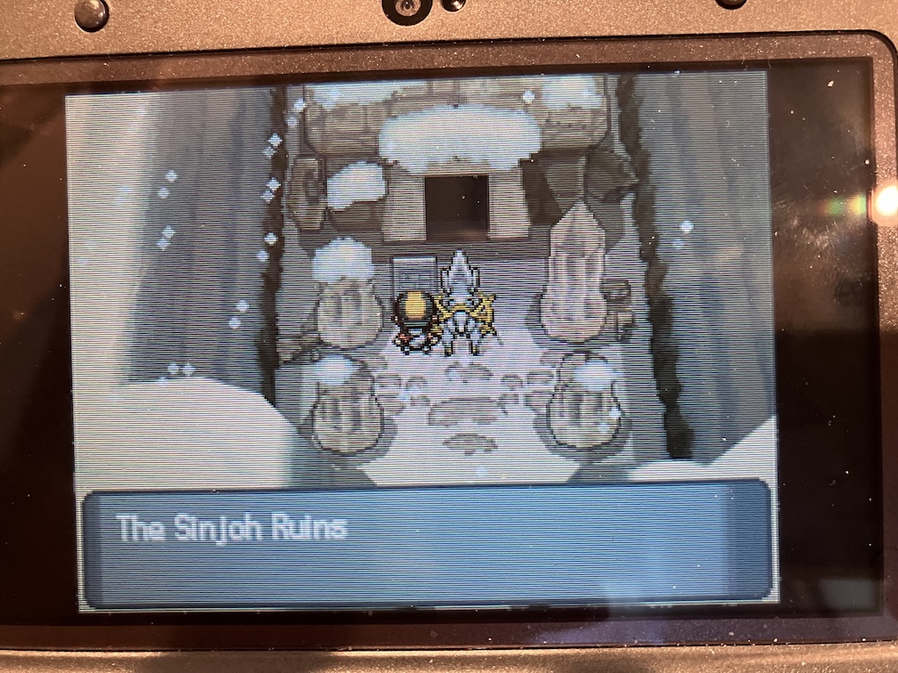 A photo of Pokemon HeartGold. The player and Arceus are standing in front of an old ruined building in a snow covered crevice. The sign reads "The Sinjoh Ruins".