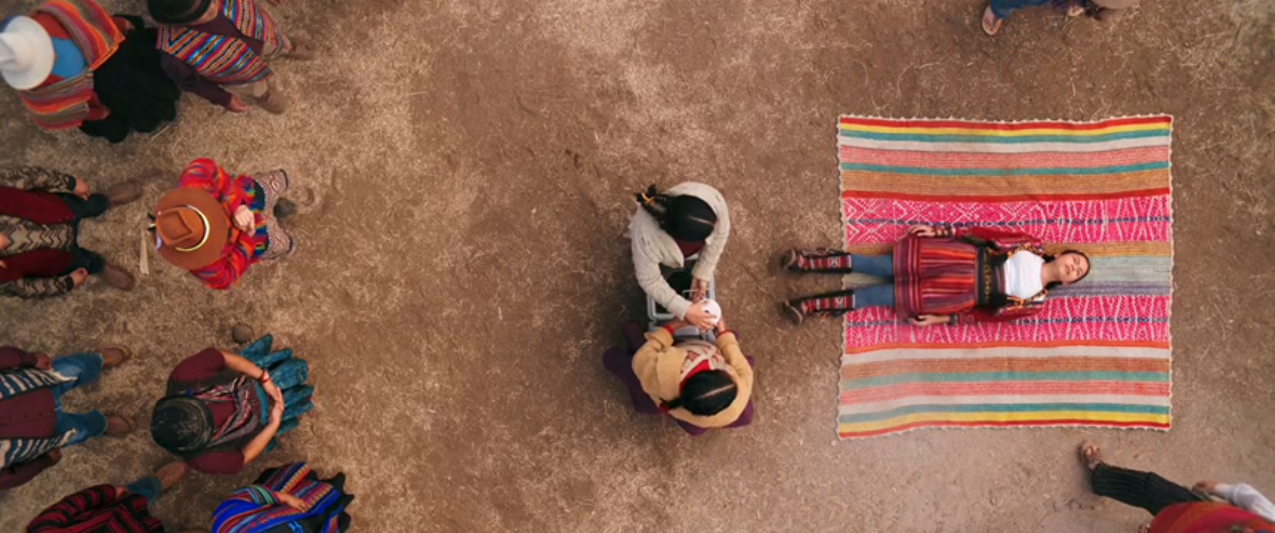 On the right, Kusimaya is lying and convulsing on a blanket. In the middle, two people stand holding a cannister. On the left, a crowd of previously-singing people watch Kusimaya.